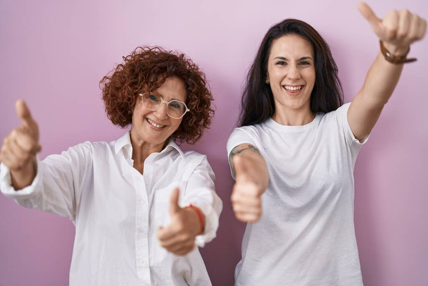 Hispánská matka a dcera na sobě ležérní bílé tričko přes růžové pozadí schvalující dělá pozitivní gesto rukou, palce nahoru s úsměvem a šťastný za úspěch. gesto vítěze.  - Fotografie, Obrázek