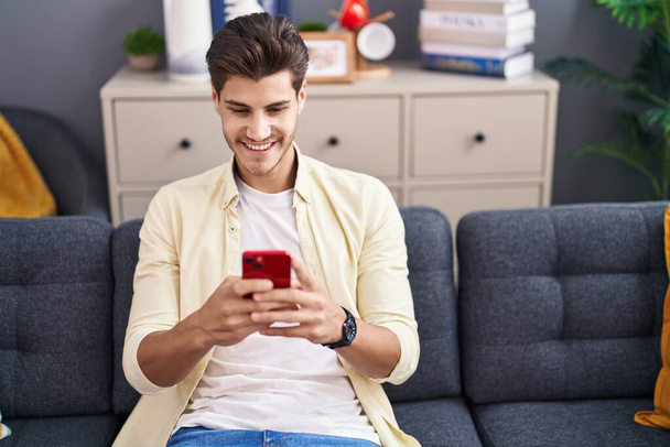Fiatal spanyol férfi használ okostelefon ül kanapén otthon - Fotó, kép