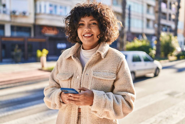 Jeune belle femme hispanique souriant confiant en utilisant smartphone à la rue - Photo, image