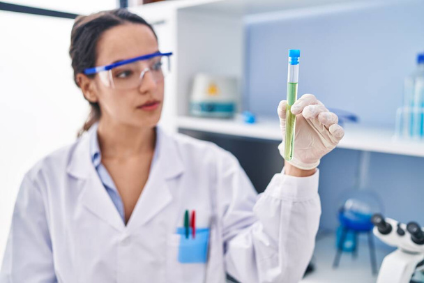 Mujer hispana joven vistiendo uniforme científico sosteniendo tubo de ensayo en laboratorio - Foto, imagen