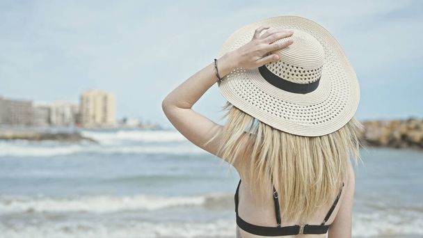Joven turista rubia con bikini y sombrero de verano de pie hacia atrás en la playa - Foto, imagen
