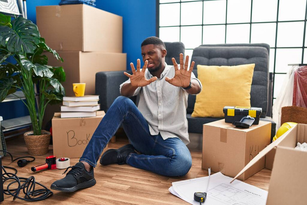 Afroamerikaner, der in seinem neuen Zuhause ängstlich und verängstigt auf dem Boden sitzt, stoppt die Geste mit den Händen und schreit vor Schreck. Panikkonzept.  - Foto, Bild