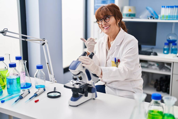 Keski-ikäinen latino nainen työskentelee tiedemies laboratoriossa osoittaa peukalo ylös puolelle hymyillen onnellinen suu auki  - Valokuva, kuva
