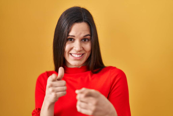 Fiatal spanyol nő áll a sárga háttér mutató ujjak kamera boldog és vicces arcát. jó energia és rezgések.  - Fotó, kép
