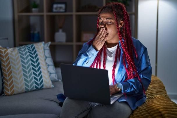 Mujer afroamericana con cabello trenzado usando computadora portátil por la noche aburrido bostezo cansado cubriendo la boca con la mano. inquietud y somnolencia.  - Foto, imagen