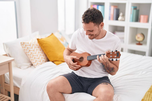 Jeune homme hispanique jouant ukulele assis sur le lit dans la chambre - Photo, image