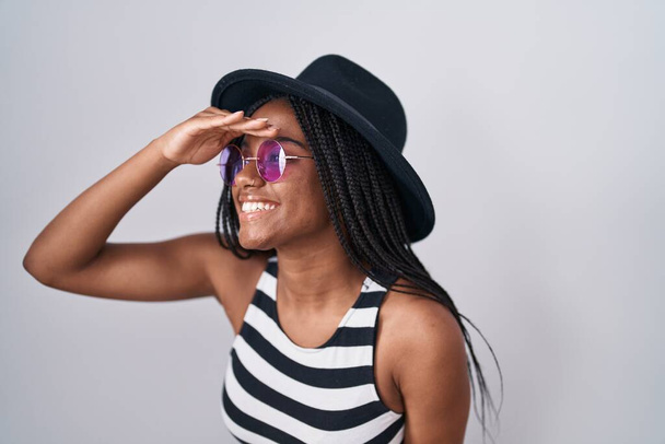 Young african american with braids wearing hat and sunglasses very happy and smiling looking far away with hand over head. searching concept.  - Photo, Image