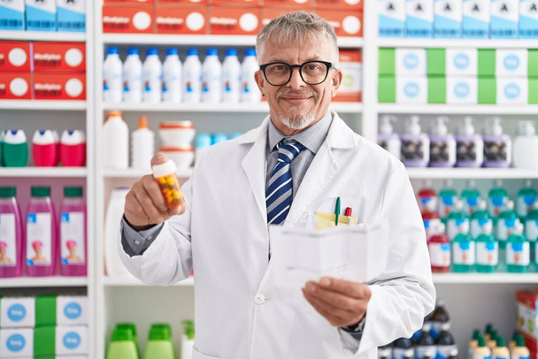 Středního věku šedovlasý muž lékárník drží pilulky čtení lahvičky předpis v laboratoři - Fotografie, Obrázek