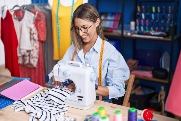 Giovane bella donna ispanica sarto sorridente fiducioso utilizzando la macchina da cucire in fabbrica di abbigliamento - Foto, immagini