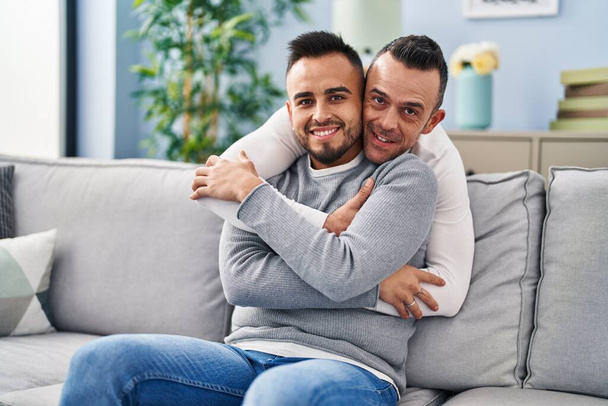 Δύο άνδρες ζευγάρι αγκαλιάζονται καθισμένοι στον καναπέ στο σπίτι - Φωτογραφία, εικόνα