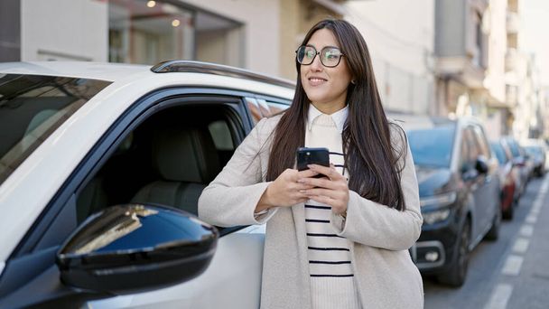 Молодая красивая латиноамериканка с помощью смартфона опирается на автомобиль на улице - Фото, изображение
