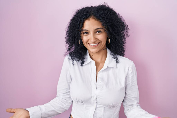 Hispanic woman with curly hair standing over pink background smiling cheerful with open arms as friendly welcome, positive and confident greetings  - Photo, Image