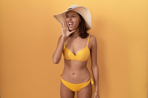 Jonge Spaanse vrouw in bikini en zomerhoed schreeuwend en schreeuwend met hand op mond. communicatieconcept.  - Foto, afbeelding