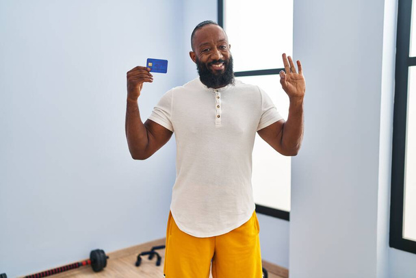 Afrykański Amerykanin w odzieży sportowej trzyma kartę kredytową robi znak ok palcami, uśmiechając przyjazny gest doskonały symbol  - Zdjęcie, obraz