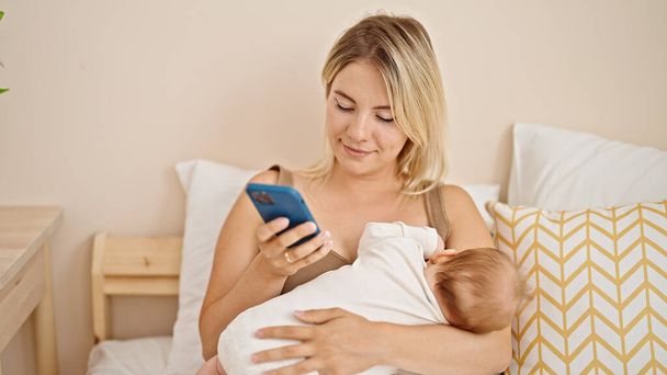 Mutter und Tochter sitzen mit Smartphone im Schlafzimmer im Bett und stillen Baby - Foto, Bild