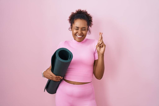 Junge hispanische Frau mit lockigem Haar hält Yogamatte über rosa Hintergrund gestikulierend den Finger gekreuzt lächelnd mit Hoffnung und geschlossenen Augen. Glück und abergläubisches Konzept.  - Foto, Bild