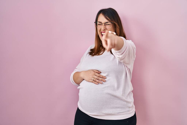 Donna incinta in piedi su sfondo rosa ridere di te, puntando il dito verso la fotocamera con mano sul corpo, espressione di vergogna  - Foto, immagini