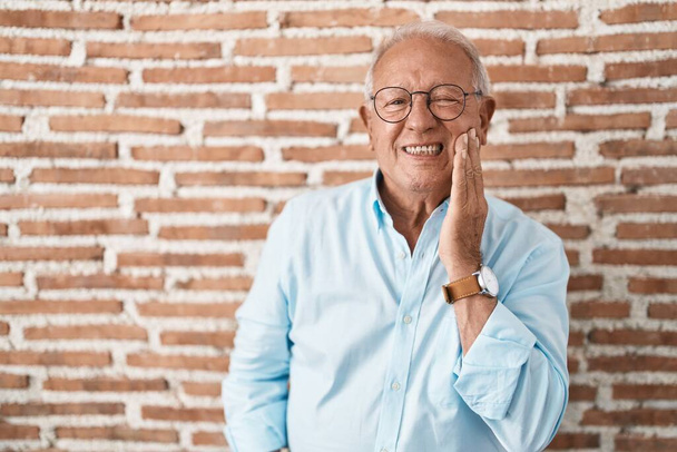 Älterer Mann mit grauen Haaren, der über Ziegelsteinen steht und den Mund mit schmerzverzerrtem Gesichtsausdruck berührt, wegen Zahnschmerzen oder Zahnkrankheiten an den Zähnen. Zahnarzt  - Foto, Bild