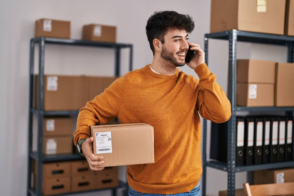 Молодий іспаномовний працівник електронної комерції розмовляє на смартфоні, тримаючи пакет в офісі
 - Фото, зображення