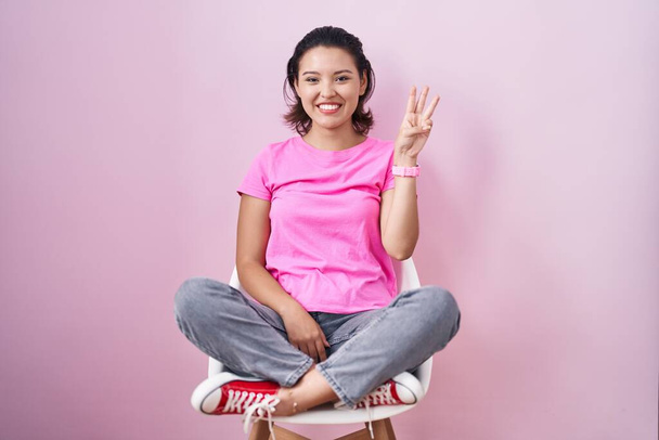 Hispanic young woman sitting on chair over pink background showing and pointing up with fingers number three while smiling confident and happy.  - 写真・画像
