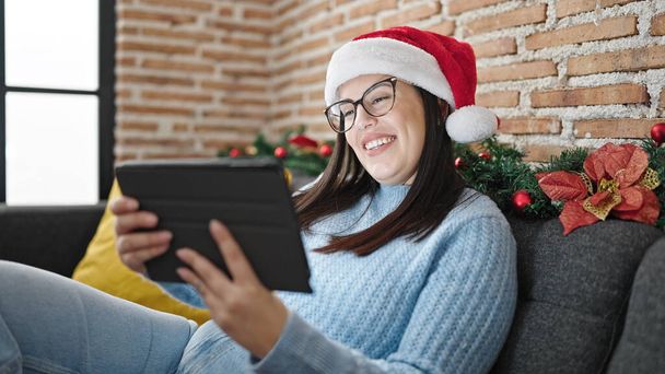 Mujer hispana joven usando touchpad sentado en el sofá con sombrero de Navidad en casa - Foto, imagen