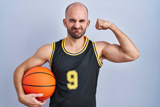 Jeune homme chauve avec barbe portant l'uniforme de basket-ball tenant ballon personne forte montrant muscle du bras, confiant et fier de la puissance  - Photo, image