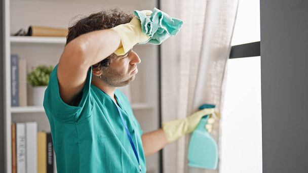 Joven hombre hispano limpiando ventana con un paño cansado en casa - Foto, Imagen