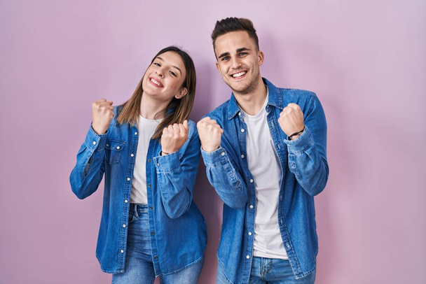 Молодая латиноамериканская пара, стоящая на розовом фоне, очень счастлива и взволнована, делая жест победителя с поднятыми руками, улыбаясь и крича об успехе. празднование концепции.  - Фото, изображение