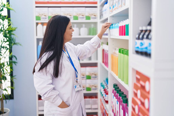 Joven hermosa mujer hispana farmacéutica sonriendo confiado sosteniendo producto en estanterías en la farmacia - Foto, Imagen