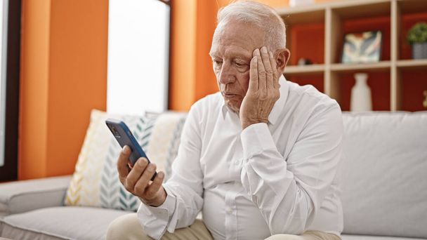 Hombre mayor de pelo gris preocupado con la mano en la cabeza usando el teléfono inteligente en casa - Foto, imagen