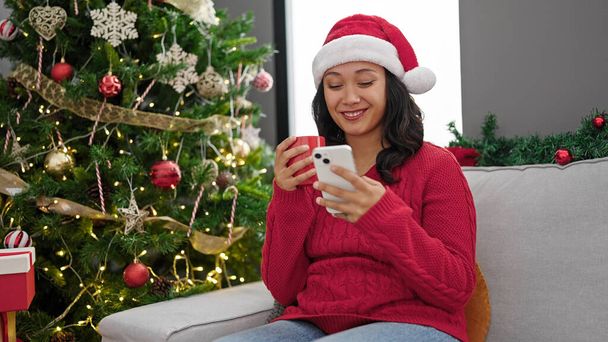 Молодая красивая латиноамериканка с помощью смартфона пьет кофе, празднуя Рождество дома - Фото, изображение