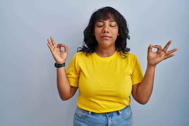 Hispanic woman standing over blue background relaxed and smiling with eyes closed doing meditation gesture with fingers. yoga concept.  - Фото, изображение