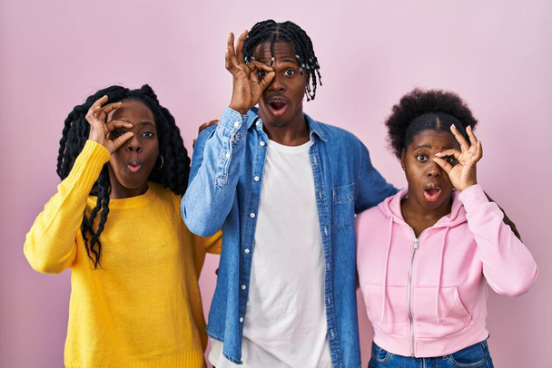 Group of three young black people standing together over pink background doing ok gesture shocked with surprised face, eye looking through fingers. unbelieving expression.  - Zdjęcie, obraz