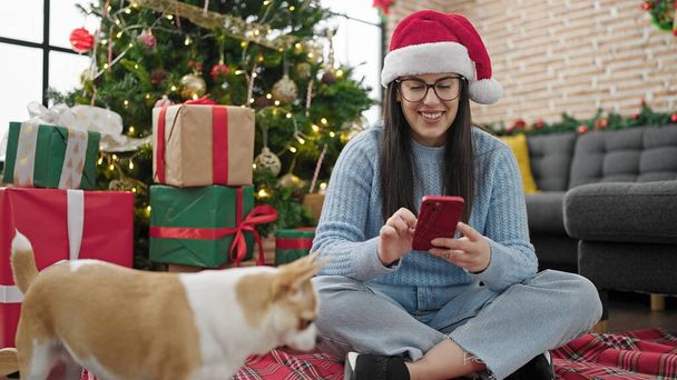 Mujer hispana joven con perro chihuahua usando teléfono inteligente sentado en el suelo por árbol de Navidad en casa - Foto, imagen