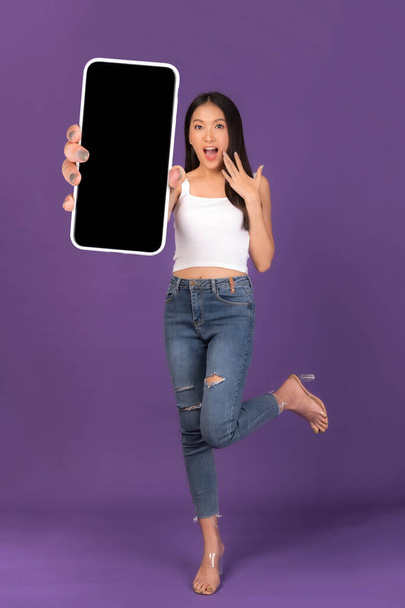 Фотографія прекрасної азіатської дівчини з повною довжиною тіла, вражена здивованою дівчиною, яка показує великий смарт-телефон з чистим екраном, білий екран ізольований на пурпуровому фоні, Mock Up Image - Фото, зображення