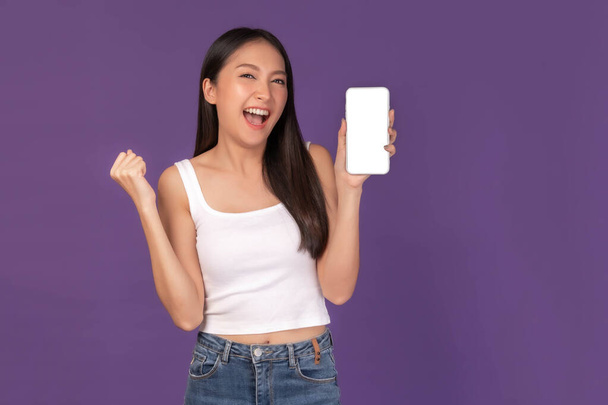 retrato de hermosa mujer joven asiática mostrando teléfono inteligente con pantalla en blanco, pantalla blanca espacio de copia vacío para banner de publicidad aislado en el fondo púrpura, imagen simulada - Foto, imagen