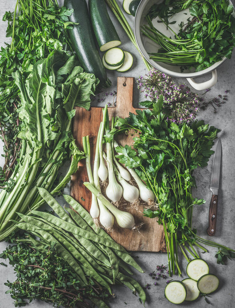 Variety of green vegetables and herbs on a rustic kitchen table with a wooden cutting board and pans for healthy cooking at home. Top view. - Foto, Imagem