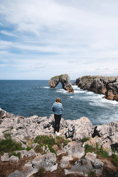 young woman observing the Castro de las Gaviotas from the cliffs of Huelga beach on the Asturian coast, near LLanes - 写真・画像