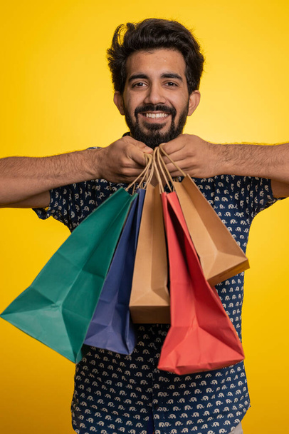 Young indian man showing shopping bags, advertising discounts, smiling looking amazed with low prices, shopping on Black Friday holidays. Shopaholic hindu guy isolated on studio yellow background - Fotó, kép