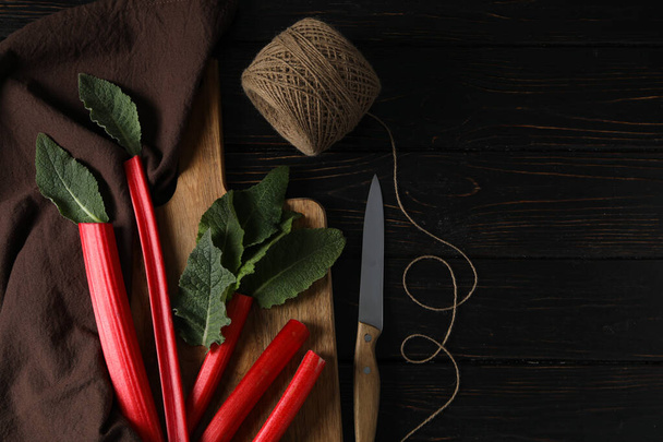 Rhubarb stalks and mint on board, brown towel, twine and knife on dark wooden background, space for text - Photo, image