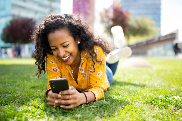 Portrait de jeune femme africaine heureuse allongée sur l'herbe et utilisant le téléphone mobile - Photo, image