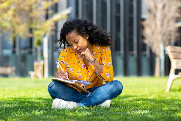 Portrait of young mixed race woman sitting on grass in park writing in book - Fotó, kép