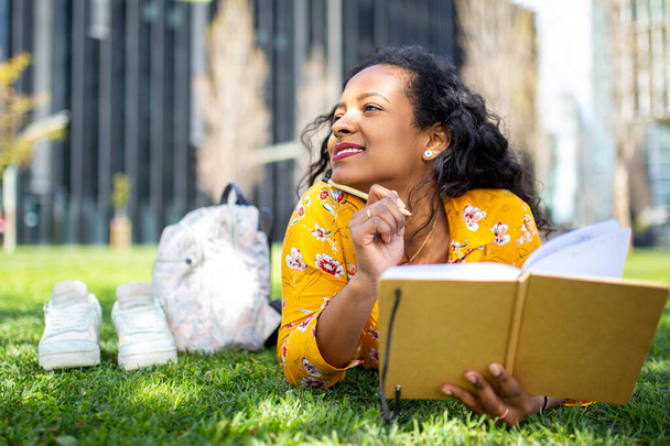 Genç Afrikalı bir kadının portresi parkta çimlere uzanmış, bir kitap yazıyor, başka tarafa bakıyor ve düşünüyor. - Fotoğraf, Görsel
