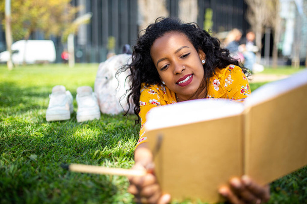 Retrato de una joven afroamericana tumbada sobre hierba en el parque escribiendo un libro y pensando - Foto, Imagen