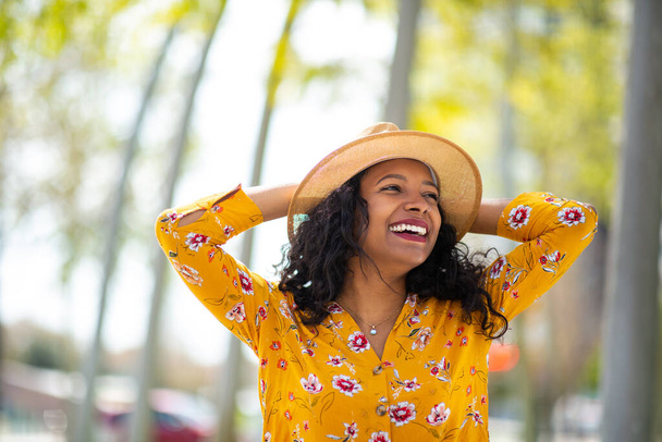 Portret van een lachende jonge Afrikaanse vrouw die buiten in een hoed staat en wegkijkt. - Foto, afbeelding