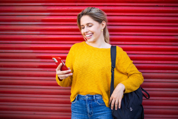 Porträt einer glücklichen kaukasischen Frau, die auf ihr Handy schaut und lächelt, während sie vor dem roten Verschluss steht - Foto, Bild