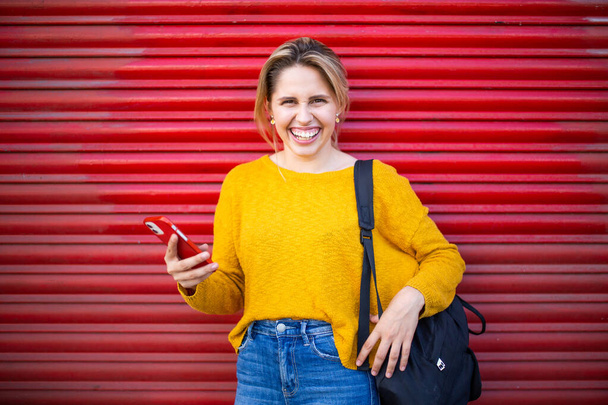 Portrait of cheerful young woman with phone and bag standing in front of red shutter - Фото, изображение