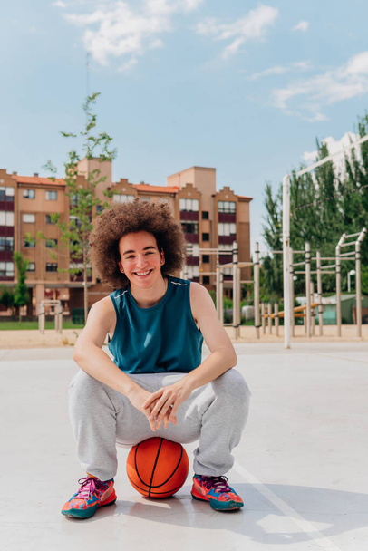Glimlachende sportieve jongen zittend op een basketbal in een stedelijk veld - Foto, afbeelding