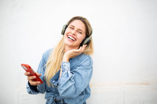 Portré vidám fiatal nő mobiltelefonnal és fülhallgatóval zenét hallgatni a fehér háttér - Fotó, kép