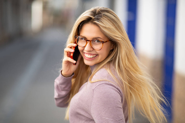 Close up portrait of beautiful young blond woman looking behind and talking on cellphone in city - Photo, Image